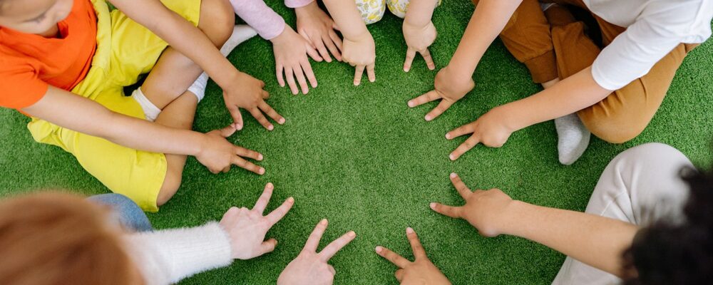 Kinderen zitten op het gras in een cirkel en hebben hun wijs- en middelvinger van een hand gespreid op de grond - Foto: Yan Krakau, pexels