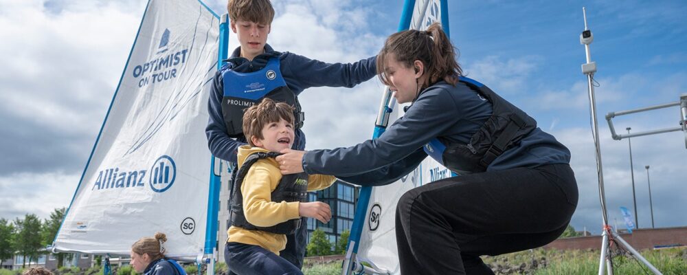 Zeilinstructrice doet een zwemvest aan bij een klein kind dat in een zeilbootje staat met een ander kindje.