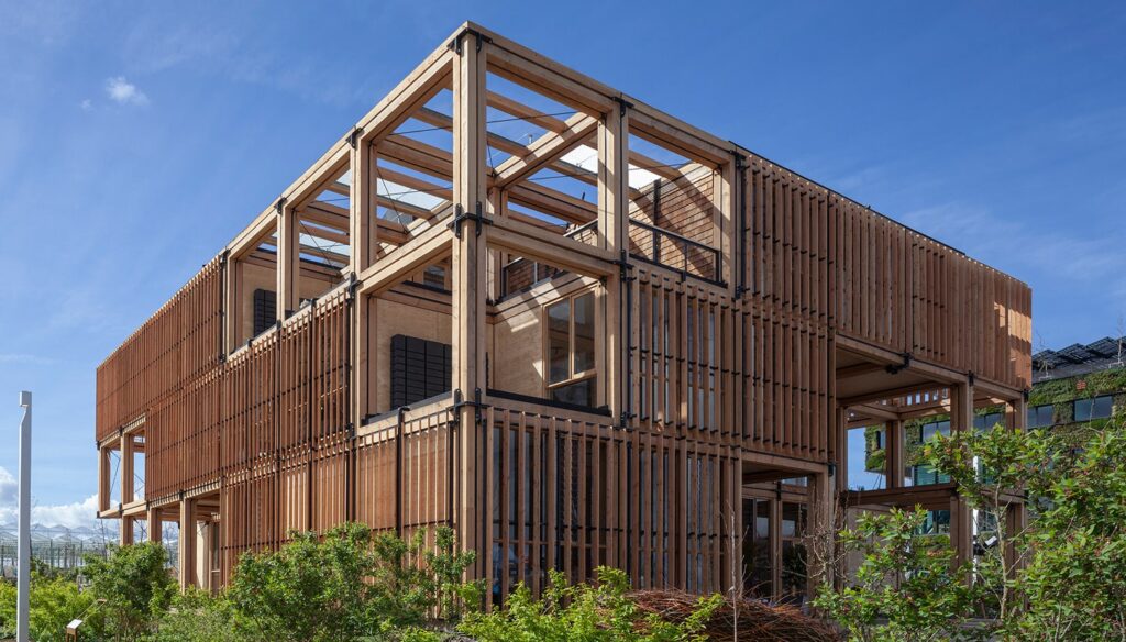 Een modern gebouw met een open houten structuur, grote ramen en omgeven door groene planten onder een blauwe lucht.