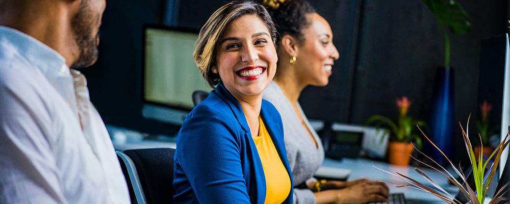 Een vrouw in een blauwe blazer en gele top glimlacht naar een collega in een modern kantoor.