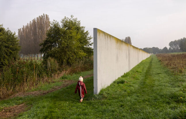 Een klein kind met een muts en rode jas loopt op een pad naast een grote witte muur, omringd door gras, bomen en een bewolkte lucht.