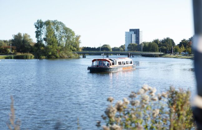 Een rondvaartboot vaart rustig over een breed kanaal, omgeven door groene bomen en een gebouw op de achtergrond.