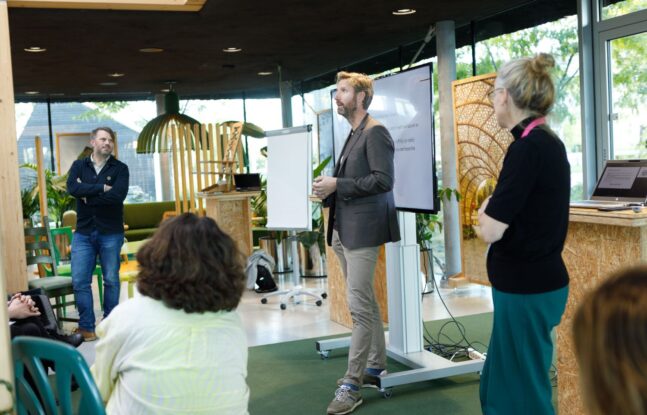 Een man staat vooraan in een zaal en geeft een presentatie, terwijl twee mensen toekijken. Achter hem is een scherm met tekst te zien.