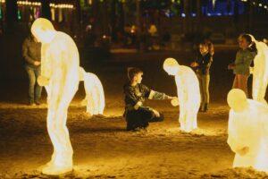 Een jongen raakt een lichtgevende, mensvormige sculptuur aan, terwijl andere kinderen toekijken op een zandige ondergrond.