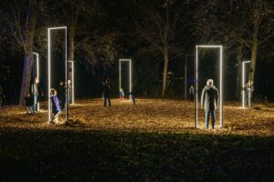 Personen staan 's avonds in een bos omringd door grote, rechthoekige lichtframes die een mysterieuze sfeer creëren.