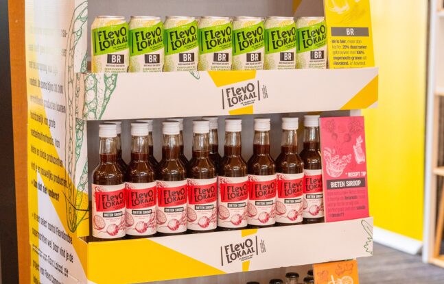 FlevoLokaal producten, waaronder blikken bier en flessen bietensiroop, gepresenteerd op een winkeldisplay met een recepttip voor bietensiroop - Foto: Calina Inc