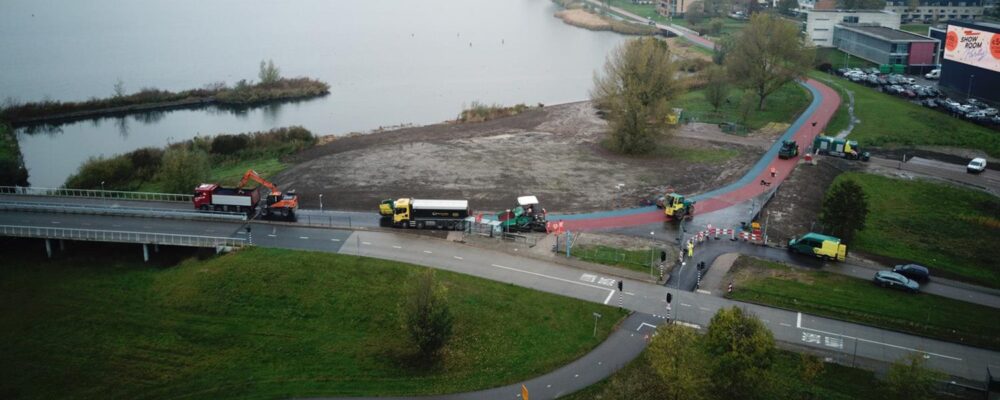 Luchtfoto van werkzaamheden aan een rood en blauw fietspad langs het Weerwater, met graafmachines en vrachtwagens op de bouwplaats.